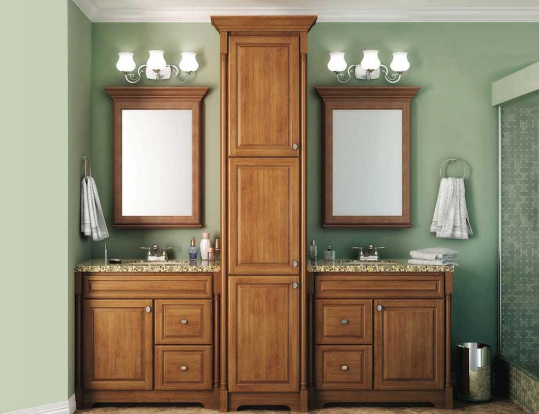 This classic bathroom features two wooden vanities separated by a tall central cabinet, each with a mirror, sink, towel ring, and three-light fixture. Green walls complement the granite counter, and a glass shower is visible on the right.