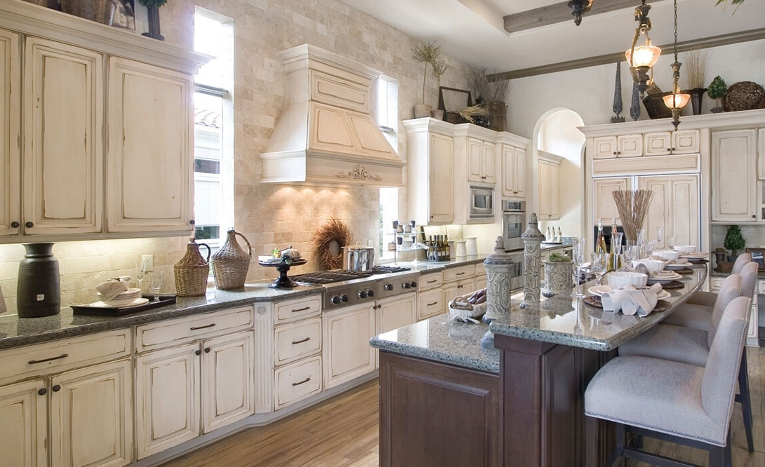 A spacious, elegant country kitchen with cream-colored cabinets, stainless steel appliances, a large island with seating, granite countertops, decorative vases, and light streaming in from tall windows.