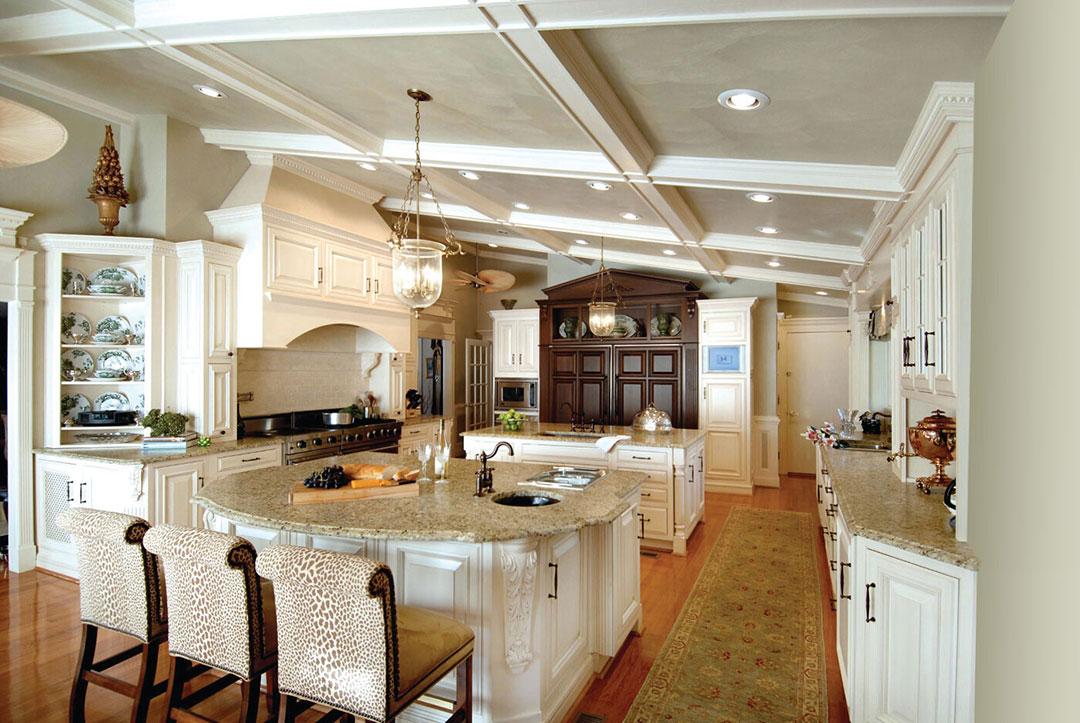Elegant, spacious kitchen with white cabinetry, granite countertops, a large island with two patterned stools, pendant lighting, wood floors, and decorative shelves—a stunning example of classic kitchens. A long rug runs along the length of the room.