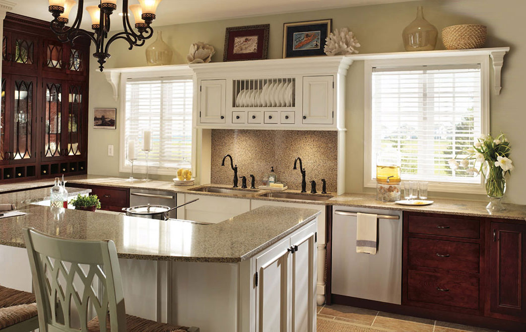 A bright, classic kitchen with white cabinets, granite countertops, a center island with chairs, two windows with blinds, a vase of flowers, and decorative items on the shelves above the sink.