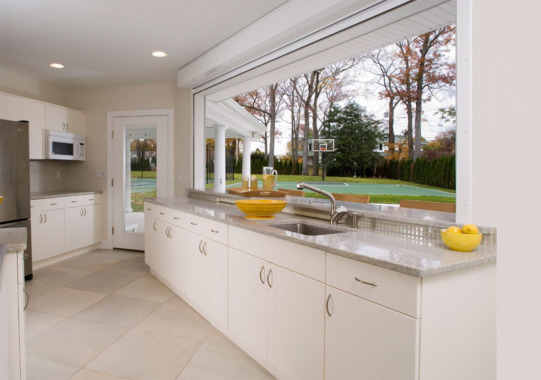 Modern kitchen with white cabinets, gray countertops, and a large open window overlooking a green backyard with trees, a basketball hoop, lawn, and stylish outdoor cabinets. A bowl of lemons and a yellow bowl sit on the counter.