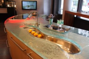 A modern kitchen island with a unique glass countertop embraces the latest trends, featuring a wavy stainless steel sink filled with lemons, a curved faucet, two blue drinks, and a tray with a metal ice bucket and glassware in a stylish interior.