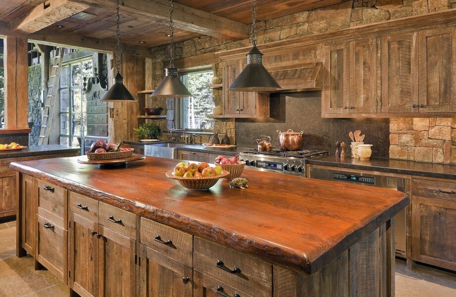 Rustic kitchen with wooden cabinets, a large island featuring a live-edge countertop, hanging black pendant lights, stone walls, and bowls of fruit. Large windows reveal lush greenery—embracing current rustic trends in kitchen design.