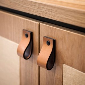 Close-up of two wooden cabinet doors with brown leather loop handles attached by black fasteners, showcasing a modern, minimalist design that reflects current trends in home decor.