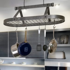 A metal kitchen rack hangs from the ceiling, holding two pots, a spatula, and a ladle. The background shows a modern kitchen with gray cabinets and stainless steel appliances, while soft spring light filters in.