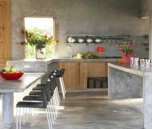 Modern kitchen embracing the latest trends with concrete countertops and floors, wooden cabinets, and black chairs around a dining table. Bright flowers, fruit bowls, and metal bowls on a wall shelf add stylish touches above the sink.