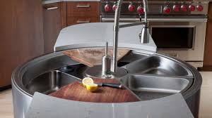 A modern circular kitchen sink workstation, reflecting the latest trends, features multiple divided stainless steel sections, a central faucet, a wooden cutting board, and half a lemon with a knife on top. Brown cabinets and a stove are in the background.