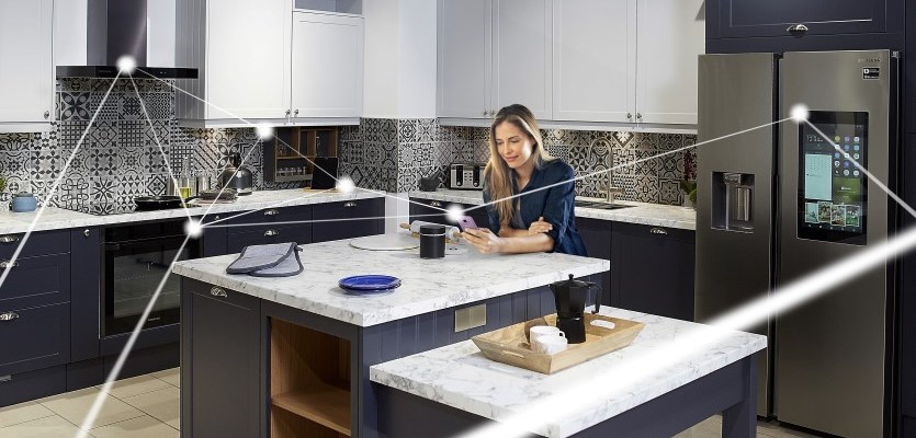 A woman sits at a kitchen island with a cup, looking at her phone. The modern kitchen features dark cabinets, patterned backsplash, and smart appliances, showcasing the latest trends with white lines representing connected smart devices.