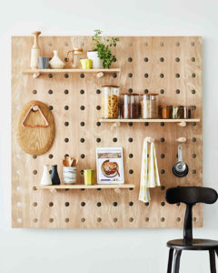 A wooden pegboard with shelves displays kitchen items, jars of pasta and grains, utensils, mugs, a cutting board, a potted plant bringing spring vibes, and a tablet with a recipe. A black chair sits in front of the pegboard.