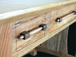Close-up of a wooden desk showing two drawers, each with a handle made of a wooden dowel and square, dark wood end pieces. The visible wood grain and natural finish reflect current trends in minimalist, stylish furniture design.