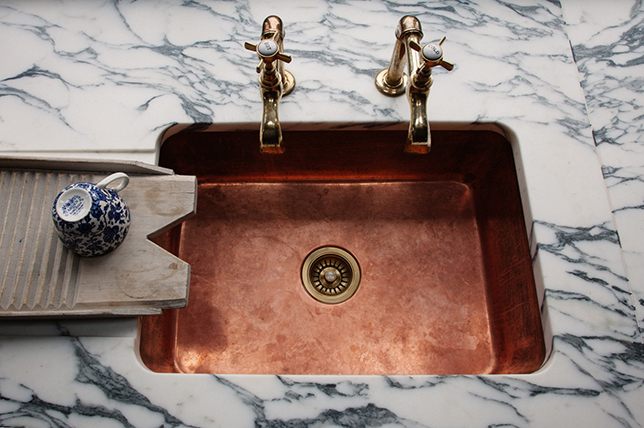 A copper kitchen sink with brass faucets set in a white marble countertop highlights current trends. A small blue and white patterned jug rests on a wooden washboard to the left of the sink.