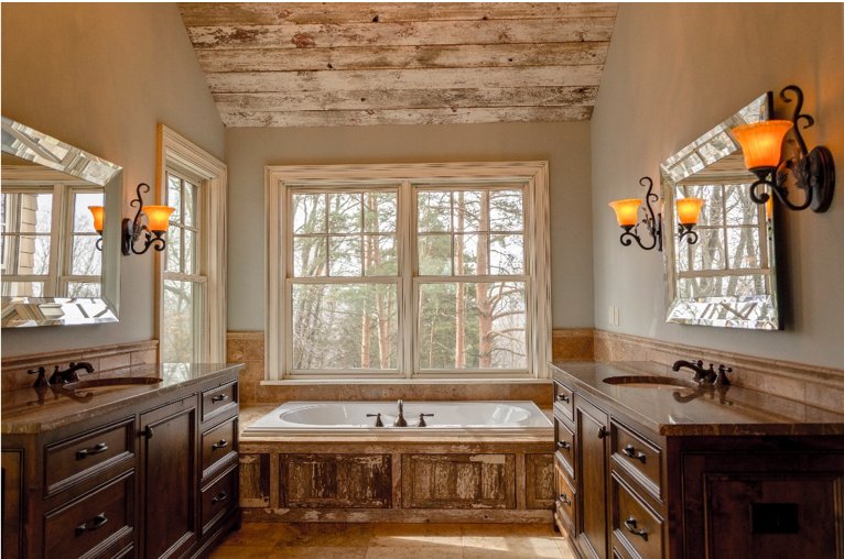 Rustic bathroom with a wood-paneled ceiling, large window, built-in bathtub, dual sinks with mirrors, dark wooden cabinets, and wall-mounted sconces with warm lighting.